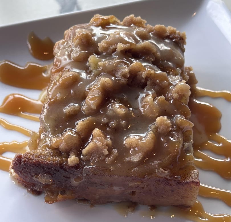 A square slice of bread pudding with a thick caramel sauce and crumble topping, drizzled with more caramel.